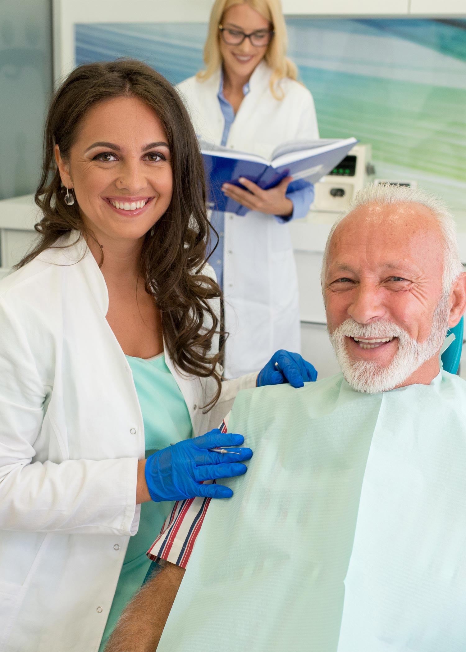 A dentist and her patient smiling into the camera as a woman looks over dental records behind them